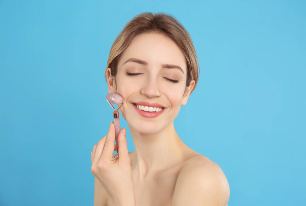 Young woman using natural rose quartz face roller on light blue background - Foto, afbeelding