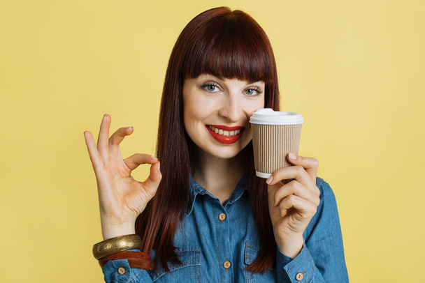 Geniet van de koffiepauze. Mooie dromerige glimlachende vrouw in jeans shirt met take away koffie kopje, poseren op geïsoleerde gele studio achtergrond en tonen oke teken, kopieer ruimte - Foto, afbeelding