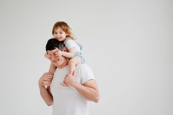 kleine dochter zit op zijn vaders schouders en sluit zijn ogen met handen - Foto, afbeelding