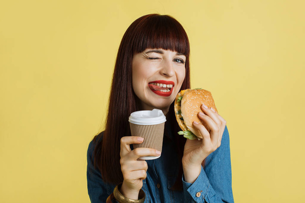 Közelkép egy vidám fiatal vonzó nő grimaszoló kamera, bemutató nyelv, gazdaság ízletes burger és elviszi a kávét, és élvezi az ebédidőt. Elszigetelve a sárga stúdió hátterében. - Fotó, kép