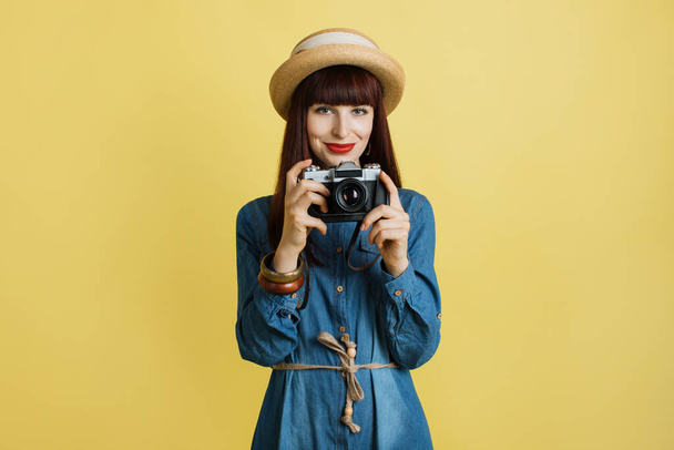 Tourism, adventures and hobby concept. Horizontal shot of smiling happy young woman in jeans dress and straw hat, holding retro photo camera, isolated on yellow background with copy space - 写真・画像