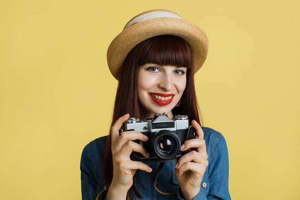Stylish red haired smiling woman photographer , wearing straw hat and denim shirt, posing with retro camera on the yellow wall background. Image with copy space - Foto, Bild