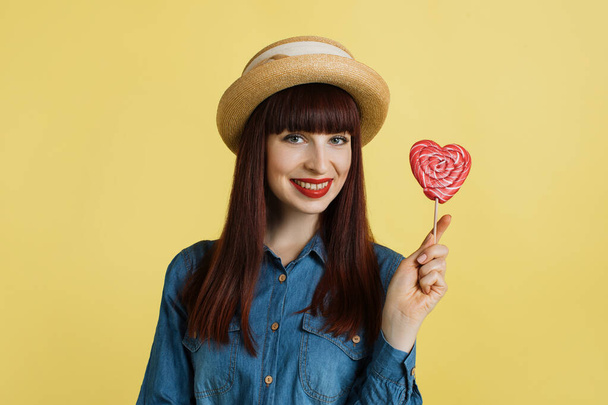Sweets, candy and positive emotions concept. Beautiful young woman in jeans shirt and hat, with red lipstick, holding a big red lollipop while standing against yellow background. - Valokuva, kuva