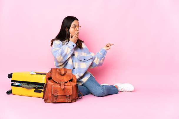 Traveler γυναίκα με μια βαλίτσα κάθεται στο πάτωμα με έκφραση έκπληξη, ενώ δείχνει πλευρά - Φωτογραφία, εικόνα