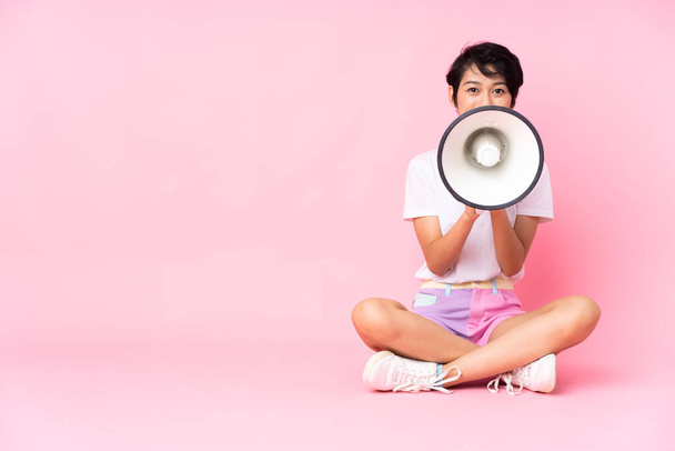 Jovem vietnamita com cabelo curto sentado no chão sobre fundo rosa isolado gritando através de um megafone - Foto, Imagem