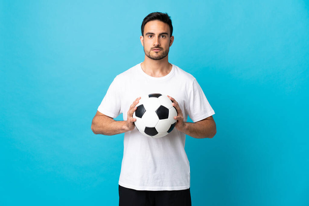 Joven hombre guapo aislado sobre fondo azul con pelota de fútbol - Foto, Imagen