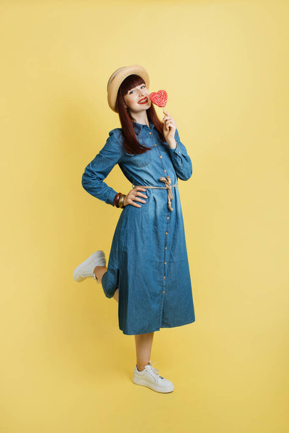 Fashion pretty girl in trendy jeans denim dress, white sneakers and straw hat, posing with one leg raised and big red lollipop, on yellow background - Fotó, kép