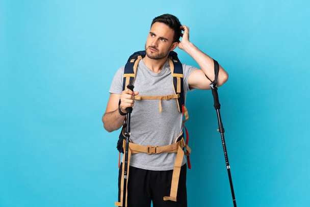 Young caucasian man with backpack and trekking poles isolated on blue background having doubts while scratching head - Foto, afbeelding