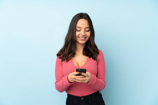 Mavi arka planda izole edilmiş genç bir ırk kadını cep telefonuyla mesaj gönderiyor. - Fotoğraf, Görsel