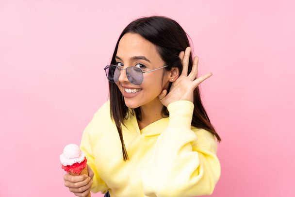 Jeune fille brune tenant une crème glacée cornet sur fond rose isolé écoutant quelque chose en mettant la main sur l'oreille - Photo, image