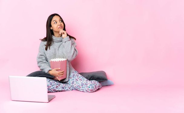 Young mixed race woman eating popcorn while watching a movie on the laptop and looking up - Φωτογραφία, εικόνα