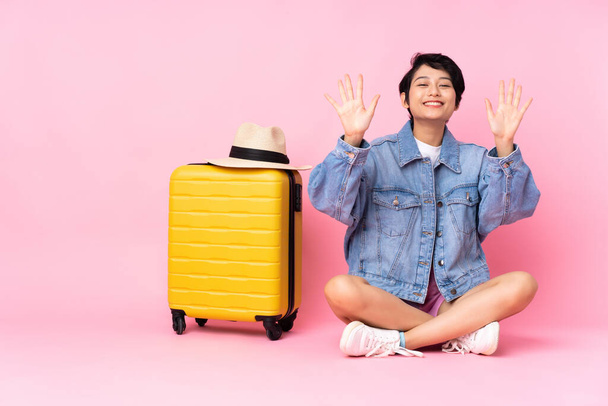 Junge reisende Vietnamesin mit Koffer sitzt auf dem Boden vor isoliertem rosa Hintergrund und zählt mit den Fingern zehn. - Foto, Bild