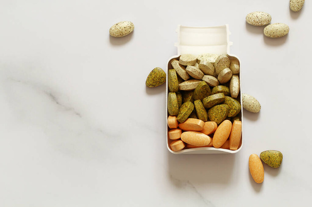 пищевая добавка в белом пластиковом контейнере, разноцветные витамины таблетки на мраморном фоне - Фото, изображение