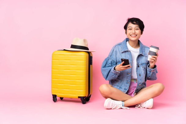 Young traveler Vietnamese woman with suitcase sitting on the floor over isolated pink background holding coffee to take away and a mobile - Fotoğraf, Görsel
