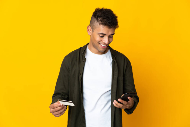 Young brazilian man isolated on yellow background buying with the mobile with a credit card - Foto, immagini