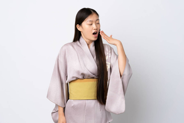 Junges chinesisches Mädchen trägt Kimono über isoliertem Hintergrund gähnt und deckt weit geöffneten Mund mit der Hand ab - Foto, Bild