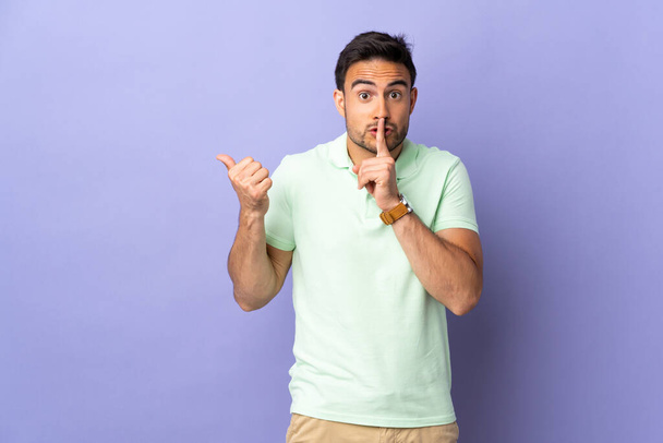 Young handsome man isolated on purple background pointing to the side and doing silence gesture - Photo, Image