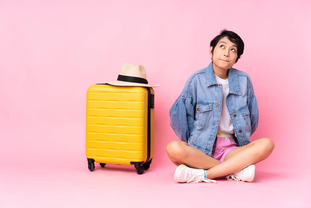 Young traveler Vietnamese woman with suitcase sitting on the floor over isolated pink background and looking up - Foto, Bild