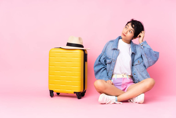 Νεαρή ταξιδιώτη Βιετναμέζα γυναίκα με βαλίτσα που κάθεται στο πάτωμα πάνω από απομονωμένο ροζ φόντο έχοντας αμφιβολίες και με συγκεχυμένη έκφραση προσώπου - Φωτογραφία, εικόνα