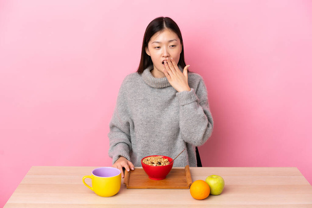 Menina chinesa nova tomando café da manhã em uma mesa bocejando e cobrindo a boca aberta larga com a mão - Foto, Imagem