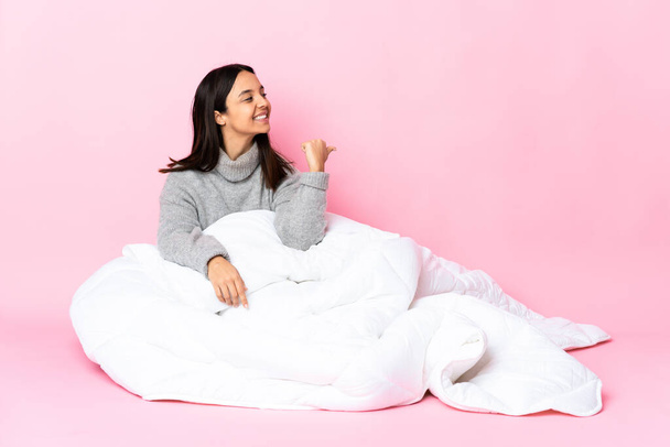 Young mixed race woman wearing pijama sitting on the floor pointing to the side to present a product - 写真・画像