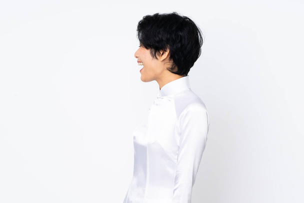 Young Vietnamese woman with short hair wearing a traditional dress over isolated white background laughing in lateral position - Foto, Imagen