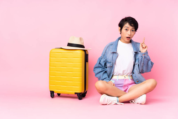 Young traveler Vietnamese woman with suitcase sitting on the floor over isolated pink background thinking an idea pointing the finger up - 写真・画像