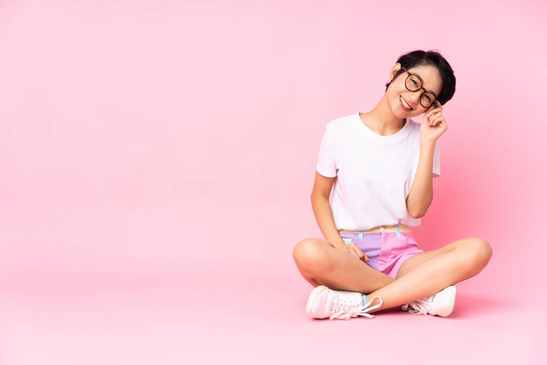 Jovem vietnamita com cabelo curto sentado no chão sobre fundo rosa isolado com óculos e feliz - Foto, Imagem