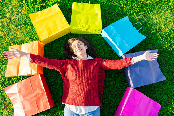 View from top of smiling girl lying on green meadow with open arms among iris colored shopping bags happy for newly purchases. Shopaholic young woman happiness buying. Modern times consumerism concept - Photo, Image