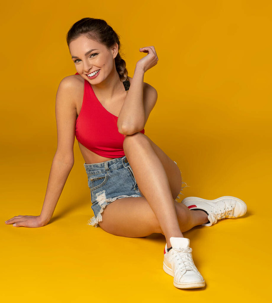 cute young model , in summer outfit , with shorts and red top , sitting on yellow background and smiling to the camera, she has snikers - Photo, Image