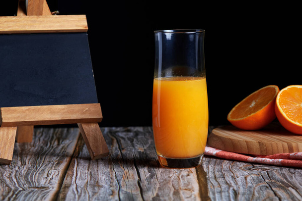 木のテーブルの上に新鮮な果物と新鮮な絞りたてのオレンジジュースのジャグ。コピースペースと黒の背景のための小さな黒板 - 写真・画像