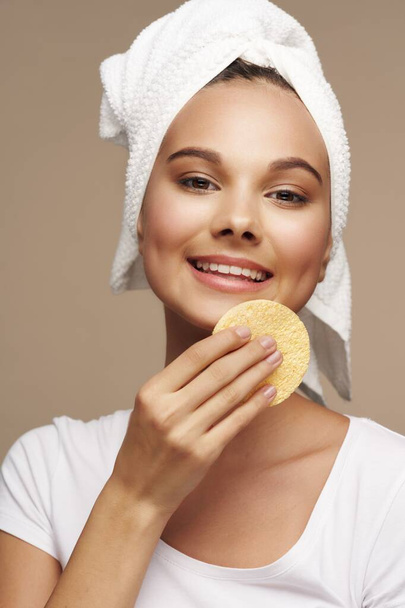 onnellinen nainen pyyhkii kasvonsa pehmeällä sienellä ja valkoisella pyyhkeellä beige tausta - Valokuva, kuva