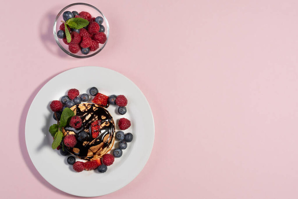 top view of homemade American pancakes with fresh blueberries, raspberries, strawberries and chocolate syrup. Healthy breakfast in the morning. Pink background - Foto, Bild