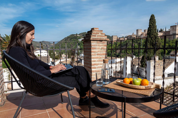 Kobieta bloger podróży pracuje na laptopie siedząc na tarasie z widokiem na twierdzę Alhambra w Granadzie, Hiszpania - Zdjęcie, obraz