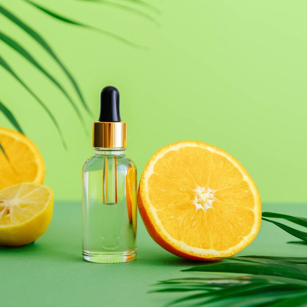 Cosmetic serum Vitamin C in glass bottle with pipette dropper. Orange essential oil with citrus ingredients Vitamin C and palm leaves on color green background. Natural skin care Cosmetics. Square - Photo, Image