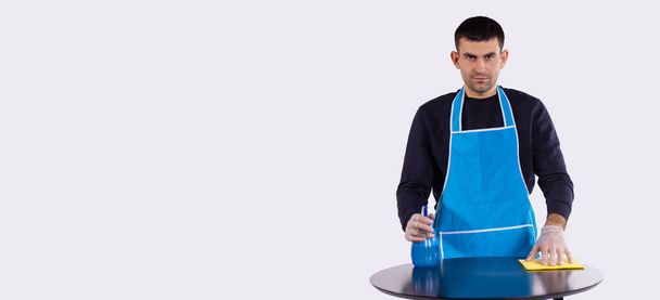 Banner, long format isolated gray background. Housekeeping and cleaning concept. Serious man wearing apron and gloves, cleaning stains off the table using blue spray and illuminating cloth - Foto, Bild