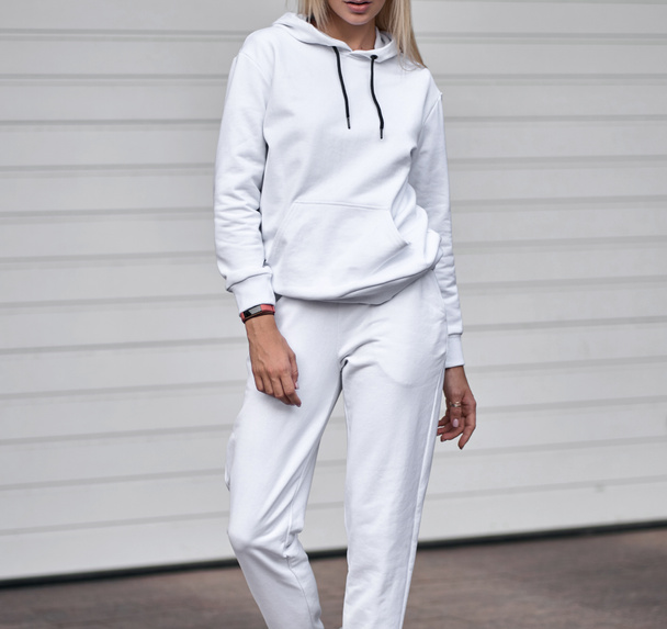 Γυναίκα στέκεται σε λευκό κοστούμι. Κομψό streetwear χωρίς λογότυπα ή φωτογραφίες σε ξανθιά κοπέλα. - Φωτογραφία, εικόνα