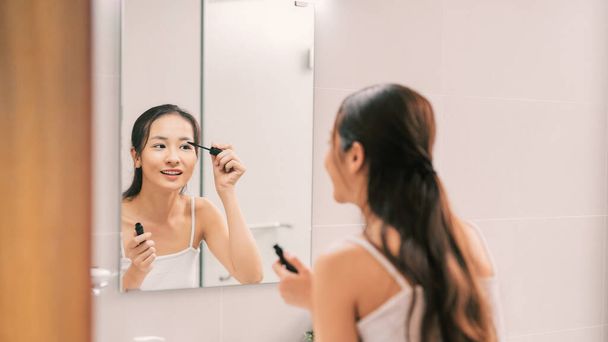 mooie jonge vrouw zetten sommige mascara op haar wimpers in de voorkant van een badkamer spiegel - Foto, afbeelding