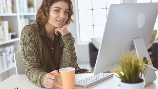 Attractive smiling woman sitting at office desk, holding a cup of coffee, she is relaxing and looking away - Zdjęcie, obraz