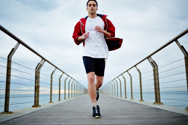 Young athlete man in white t-shirt and sports a red windbreaker runs beach pier - Φωτογραφία, εικόνα