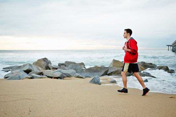 Young athlete man in white t-shirt and sports a red windbreaker runs along the beach enjoying the beautiful sea - Foto, Imagem
