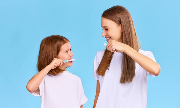 Wesoła rodzina: młoda kobieta i dziewczyna ze szczoteczkami do zębów i pastą do zębów uśmiechające się i patrząc na siebie podczas porannej rutyny higienicznej na niebieskim tle - Zdjęcie, obraz