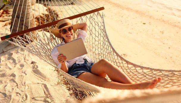 Jonge gelukkige succesvolle vrouw, lachende vrouwelijke freelancer in strohoed en zonnebril met behulp van digitale tablet, terwijl liggen en ontspannen in de hangmat op tropische zandstrand op hete zonnige dag. Afstandswerk - Foto, afbeelding