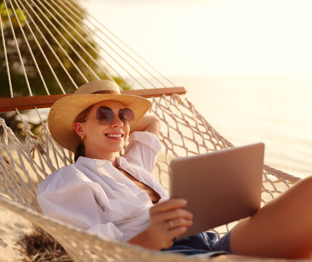 Jovem feliz mulher de sucesso, sorrindo freelancer feminino em chapéu de palha e óculos de sol usando tablet digital enquanto deitado e relaxante na rede na praia tropical de areia no dia ensolarado quente. Trabalho à distância - Foto, Imagem