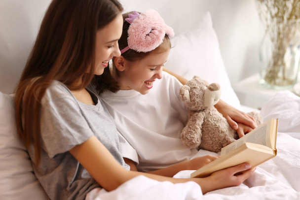 Cheerful mother hugging daughter while resting on bed and reading book together in weekend morning at home - Fotoğraf, Görsel