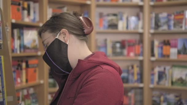 Femme caucasienne portant des lunettes et un masque contre le virus achète un livre dans un magasin - Séquence, vidéo