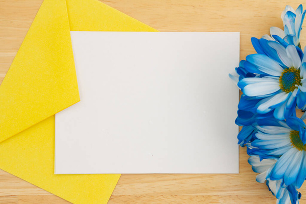 木製の机の上に青いデイジーと黄色の封筒で空白の白いグリーティングカード - 写真・画像