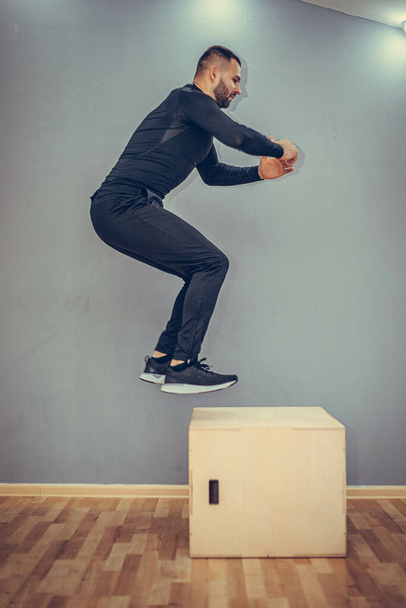 Man jumping onto the box on gray wall background. Fit athletic man jumping up to plyometric wood box, home workout exercise concept. - Foto, Imagem