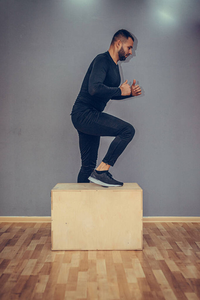 Man jumping onto the box on gray wall background. Fit athletic man jumping up to plyometric wood box, home workout exercise concept. - Photo, Image