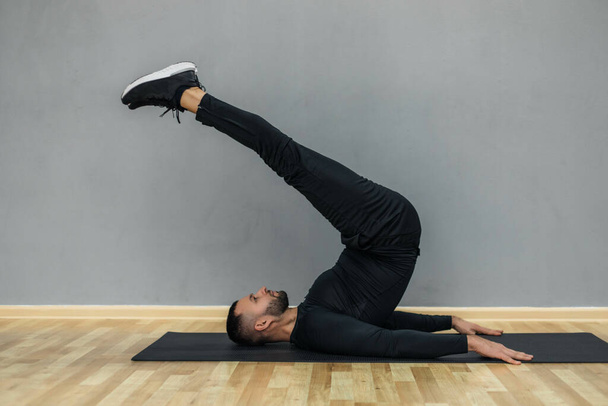 Joven hombre de fitness confiado haciendo ejercicio en una alfombra de fitness en el interior, sobre fondo de pared gris. - Foto, imagen